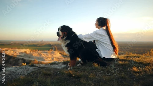 Bernese Mountain Dog (Bernese Mountain Dog). Happy young woman listening to music in headphones while walking her dog. Loving owner sitting on green lawn and playing with her big fluffy dog ​