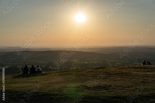 A group of people admiring the beauty of nature at sunrise, watching as the sun rises above a distant horizon over a picturesque hill. View from British camp Malvern Hills