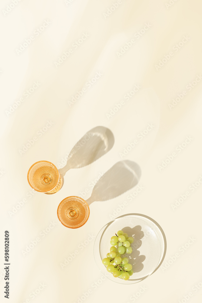 Two glasses of white wine and green grape on table, minimal summer party, shadows at sunlight, modern colored glasses on color background with blurred glares lights, copy space, top view