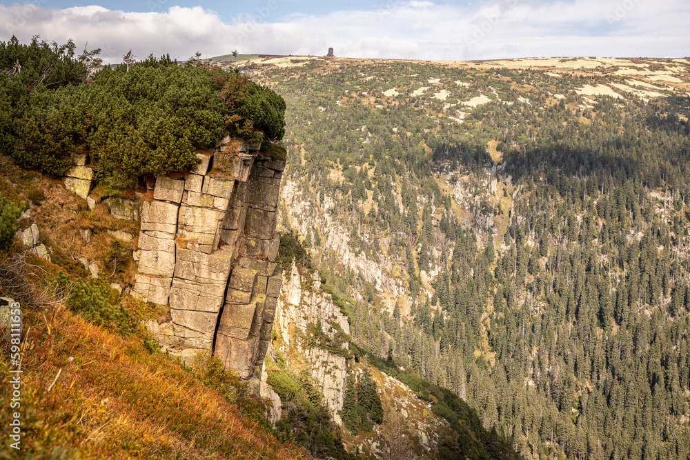 An angular mountain cliff overgrown with dwarf mountain pines. In the background a steep rocky mountain slope covered with conifers. A stone building at the top. Krkonoše National Park, Czechia.