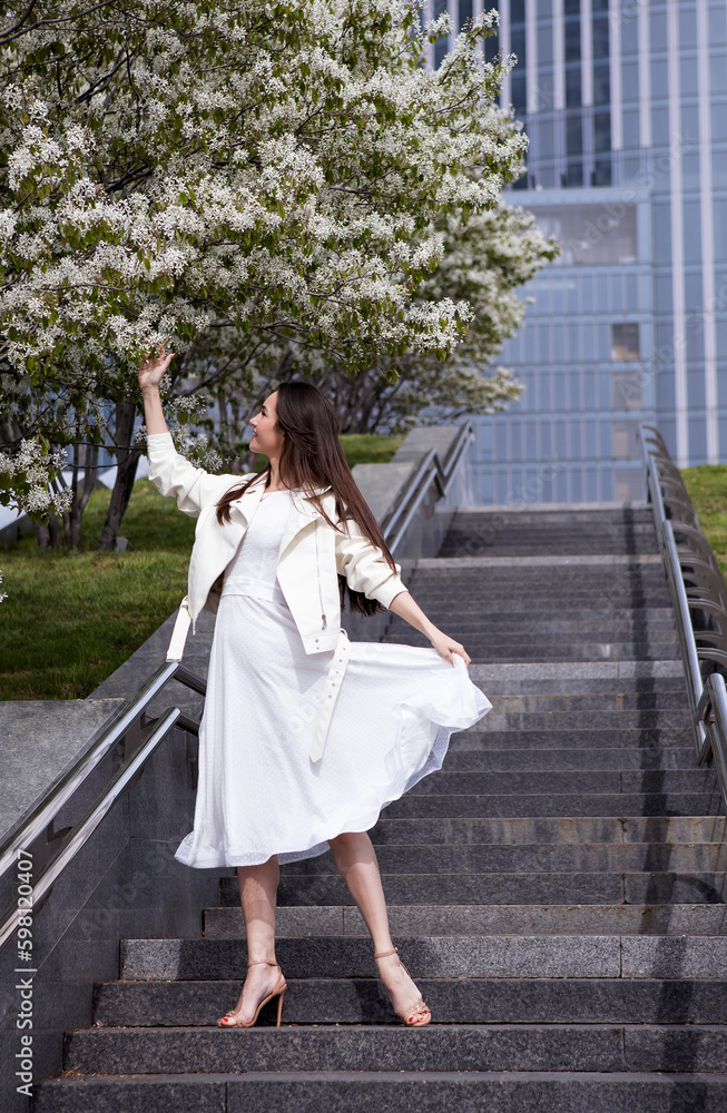 girl in a white dress stands on the stairs in a big city next to flowering trees in spring