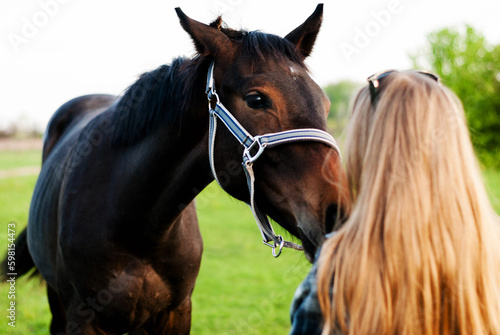 A brown horse in a bridle next to a blond woman. Farm lands. Equestrian sports school. Caring for domestic animals. Veterinary services. Race horses.