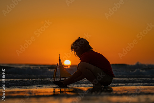 Kid playing with toy sailboat on sea beach at the summer sunset time. Sunset silhouette of child boy dreaming on cruise. Dreaming to travel. kid playing with toy sailing boat. Children day.