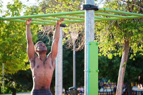 Young fit shirtless black man exercising on monkey bars outdoors on sunny summer day. Fitness and sport lifestyle.