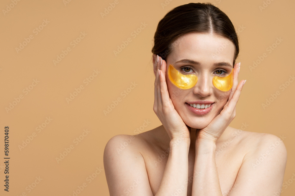 Closeup portrait of beautiful smiling caucasian woman using golden under eye patches looking at camera, isolated on beige background. Skin care, face mask, natural beauty concept Closeup portrait of beautiful smiling caucasian woman using golden under eye patches looking at camera, isolated on beige background. Skin care, face mask, natural beauty concept