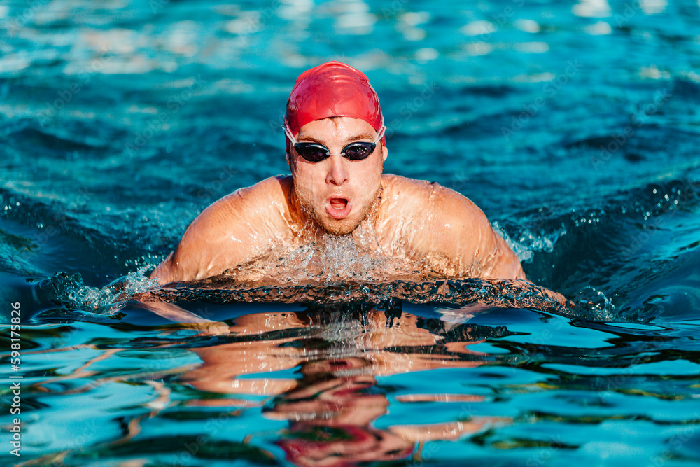 Swimming man - male swimmer swimming breaststroke. Portrait of man doing breast stroke swimming in pool outside wearing red swimming cap and swim goggles. Fit athlete training.