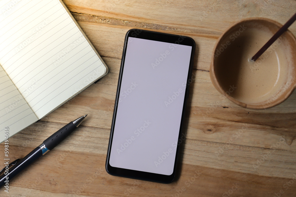 Blank screen mobile phone with empty notebook, pen, and a cup of coffee on wooden table during the day. Office work concept. Flat lay images Blank screen mobile phone with empty notebook, pen, and a cup of coffee on wooden table during the day. Office work concept. Flat lay images