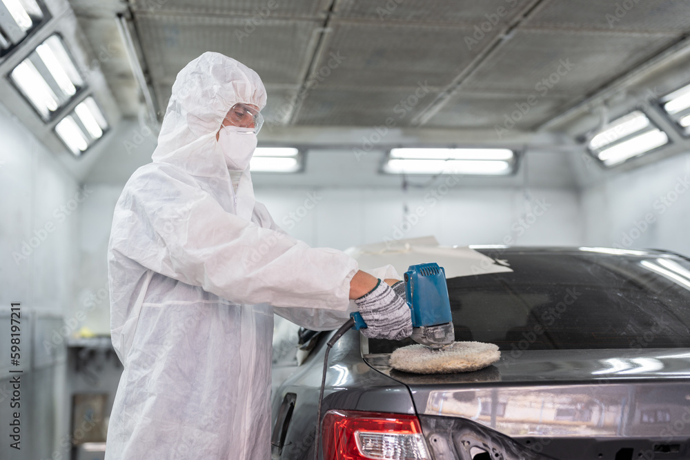 Painter man in protective workwear using a polishing machine polish car body after painting finished in the chamber at auto car garage service.