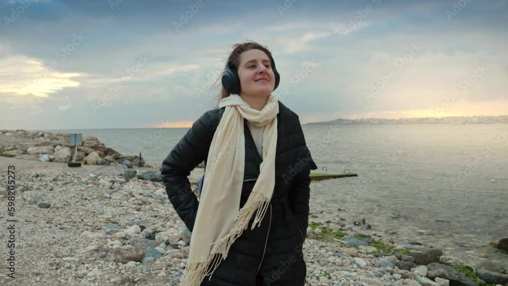 A cheerful young woman strolling along the rocky beach, headphones on and music in her ears, enjoying the cold, windy day by the sea. An ideal concept for travel, journey, and the pleasures of music A cheerful young woman strolling along the rocky beach, headphones on and music in her ears, enjoying the cold, windy day by the sea. An ideal concept for travel, journey, and the pleasures of music
