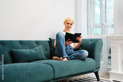 A middle aged woman with blond hair is relaxing on a sofa in her living room, reading a novel © boomeart
