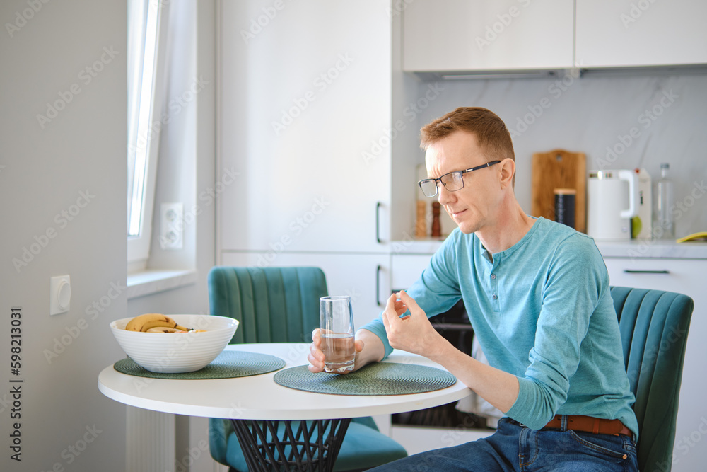 Middle aged man in the kitchen table and holding a pill and a glass of water in hands Middle aged man in the kitchen table and holding a pill and a glass of water in hands