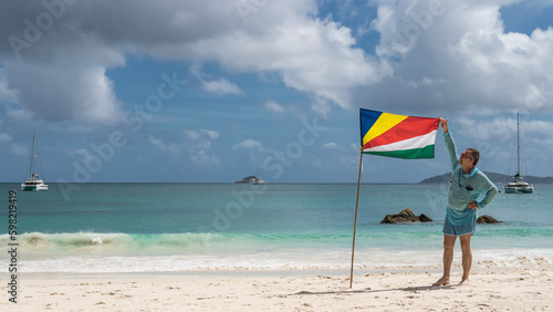 A man stands on a white sandy beach next to the flag of the Seychelles, holding a cloth around the corner. The waves of the turquoise ocean are foaming. Boulders in the water. Yachts in the distance