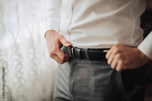 Details. A man in a white shirt and gray pants adjusts his stylish belt. Masculine style. Fashion. Business © Vasil