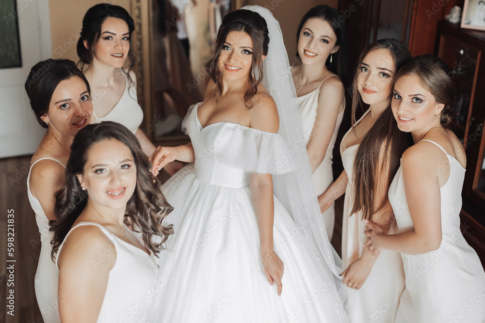 Portrait of the bride with her friends. The bride in a voluminous white dress and her beautiful friends in white evening dresses are posing. Brunettes. Open shoulders. Morning of the bride. Portrait of the bride with her friends. The bride in a voluminous white dress and her beautiful friends in white evening dresses are posing. Brunettes. Open shoulders. Morning of the bride.