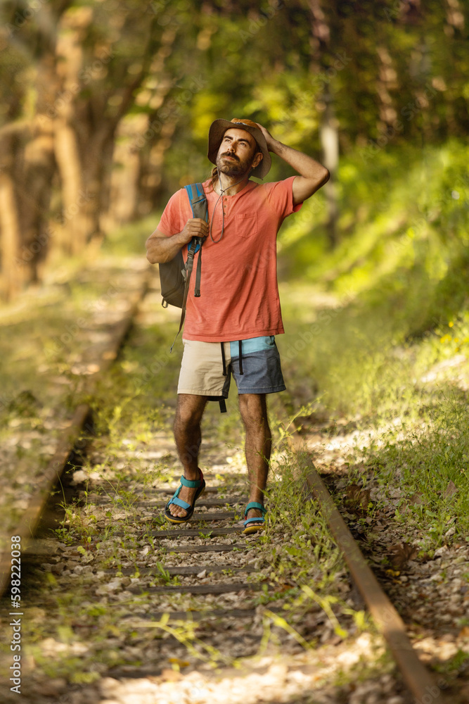 Backpacker man, looking off camera, walking along train tracks, observing Portugal's lush green forest in summertime. Wearing hat, pink t-shirt and short with sandals. Sunbeams.