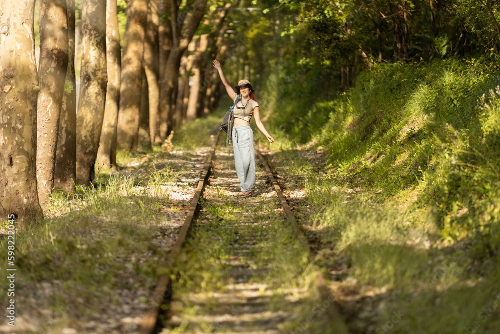 Backpacker woman, smiling, looking at camera, waving, walking along train tracks, observing Portugal's lush green forest in summertime. Wearing hat, with sandals. Sunbeams.