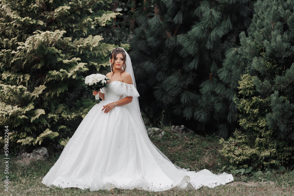 The bride in a voluminous white dress and a long veil stands on a background of green conifers with a bouquet of white roses, holding her dress. Portrait of the bride. Beautiful makeup and hair.