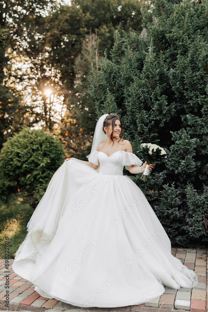 portrait of the bride in nature. A brunette bride in a voluminous dress and veil, standing, posing with a bouquet against the background of a forest and a wooden hut. A beautiful ray of the sun. portrait of the bride in nature. A brunette bride in a voluminous dress and veil, standing, posing with a bouquet against the background of a forest and a wooden hut. A beautiful ray of the sun.