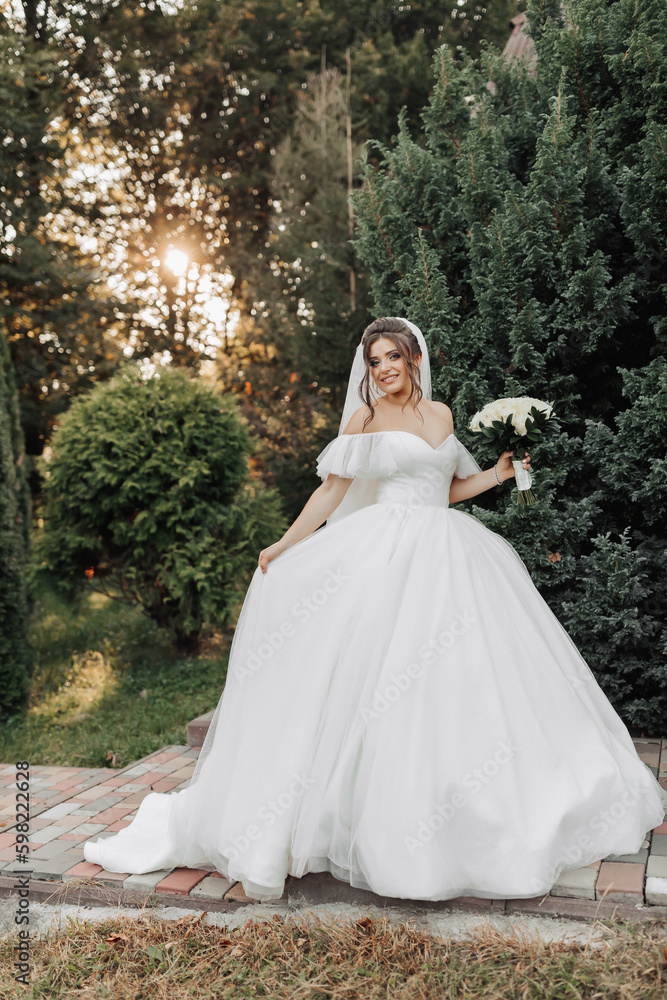 portrait of the bride in nature. A brunette bride in a voluminous dress and veil, standing, posing with a bouquet against the background of a forest and a wooden hut. A beautiful ray of the sun.