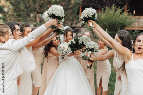 Wedding photo in nature. A brunette bride in a white long dress and her friends in nude dresses are standing against the background of trees, smiling, holding up their gypsophila bouquets. Young women