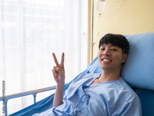 Asian patient male smile and show V sign finger while lie down on bed and recovery body from fever until healthy after that in the clinic