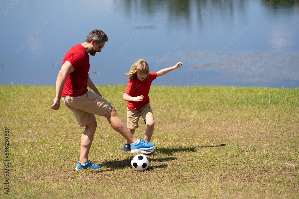Obraz premium Dad and son playing football together on green meadow. Father and son having fun on football pitch. Dad with kid are having fun and playing football on grass outdoor. Friendly family. Fathers day.