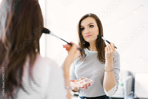 Beautiful girl puts blush on her face with a makeup brush while standing at home in a room and looking in the mirror. Caucasian girl in a striped shirt does her own makeup