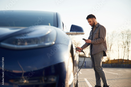 Holding the charger. Man is standing near his electric car outdoors