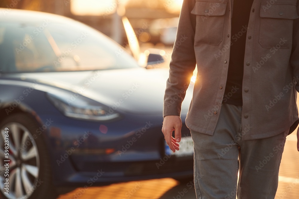Bright sunlight. Man is standing near his electric car outdoors Bright sunlight. Man is standing near his electric car outdoors