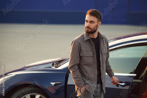 Blue colored building behind. Man is standing near his electric car outdoors