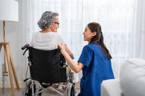 Caucasian senior woman sit on wheelchair with young Asian nurse at home. 