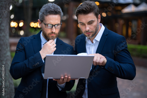 Two handsome young businessmen in classic suits using laptop. Businessmen discussing outdoor. Two business people talk project strategy. Two american businessmen in suits discuss business.