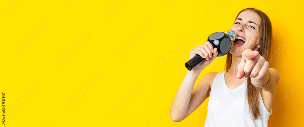 Singing young woman holding microphone pointing finger forward on yellow background. Banner Singing young woman holding microphone pointing finger forward on yellow background. Banner