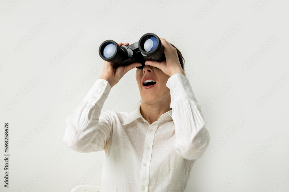 Surprised young woman in a white shirt looks through binoculars on a white background Surprised young woman in a white shirt looks through binoculars on a white background