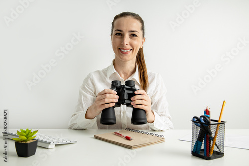 Smiling young woman sitting in the office at her desk holding binoculars