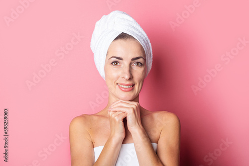 smiling young woman with a towel on her head holding her hands under her chin on a pink background.