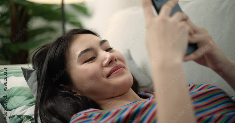 Dolly shot, Close up face of young pretty woman smile with happiness while chatting on smartphone, she lying on comfortable sofa in living room at home a small cushion under her head Dolly shot, Close up face of young pretty woman smile with happiness while chatting on smartphone, she lying on comfortable sofa in living room at home a small cushion under her head