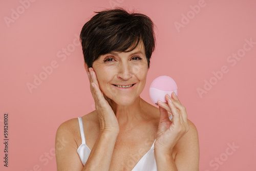 Senior woman showing cleansing facial brush isolated over pink background