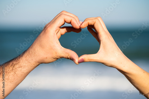 Male and female hands forming a heart shape against a sea landscape