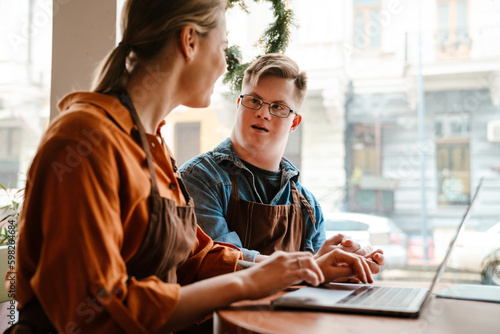 Woman using laptop while training man with down syndrome to work in cafe photo