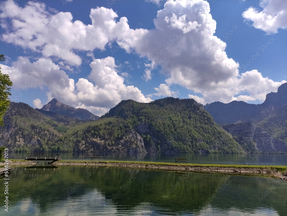 Sunny day on a river shore. Spring weater. Mountains. Blue sky and clouds. Lush green grass. Mountain and lake. Blue spring sky and white clouds.  Traunsee, Austria. Mountain lake. Reflection on water