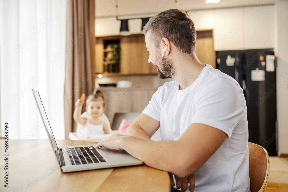 A happy father is working from home on a laptop and talking to his daughter while babysitting her.