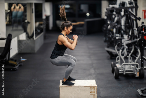 A sportswoman in shape is jumping on a box in a gym and doing cardio training.