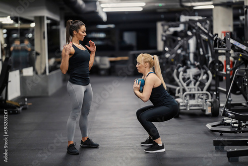 A female personal trainer is showing exercises to a sportswoman who is squatting and holding kettlebell in a gym.