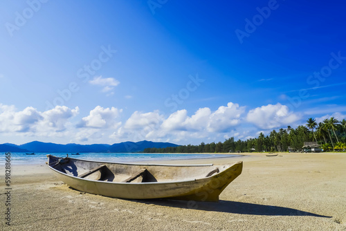 Small Indonesian traditional wooden ship or Jongkong lying on the Tropical beach with coconut trees, traditional house, and blue sky with clouds on Sunny day. © Rizky