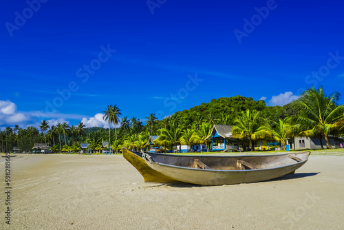 Small Indonesian traditional wooden ship or Jongkong lying on the Tropical beach with coconut trees, traditional house, and blue sky with clouds on Sunny day.