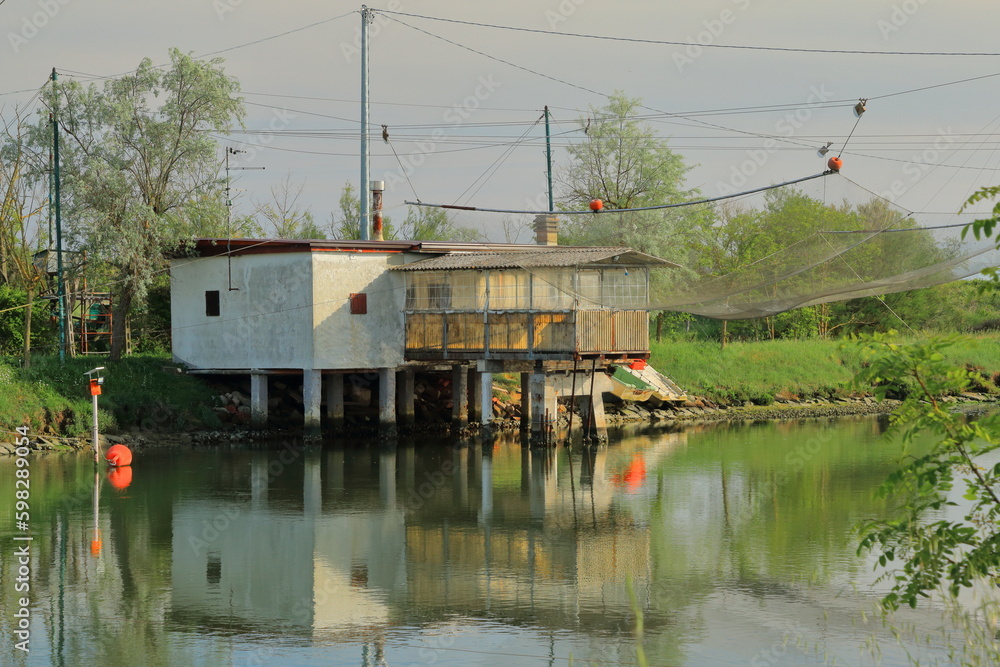 An old white fishing hut is reflected in the water of the Po Delta in Italy on a May afternoon. An old white fishing hut is reflected in the water of the Po Delta in Italy on a May afternoon.