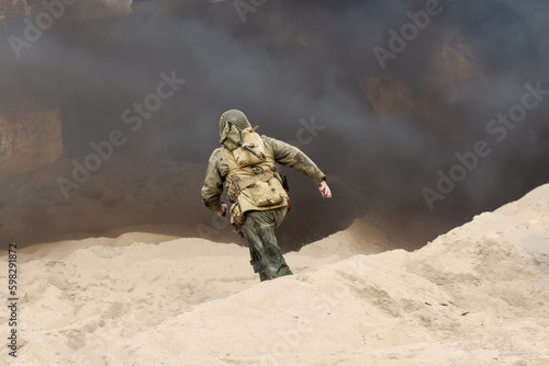 Historical reconstruction. An American infantry soldier from the World War II runs on the beach between smoke and dust. View from the back. Hel, Poland 