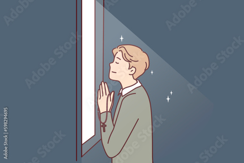Religious man prays standing at window that emits light and holds chain with christian crucifix in hands. Believing guy prays turning to god is adherent of orthodox or catholic faith Generative AI