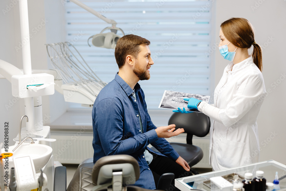 Young attractive man visiting dentist, sitting in dental chair at modern light clinic. Young woman dentist holding x ray image. Young attractive man visiting dentist, sitting in dental chair at modern light clinic. Young woman dentist holding x ray image.
